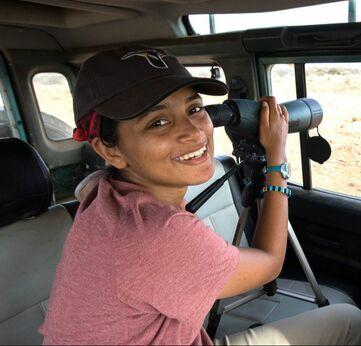 A woman with binoculars, in a vehicle, turning and smiling at camera
